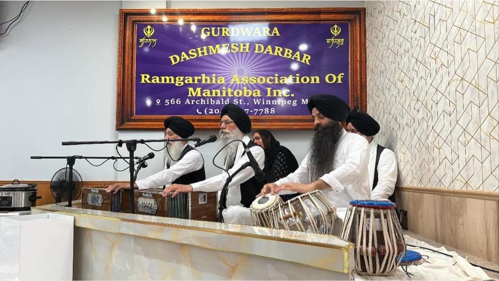 A kirtan jatha playing harmoniums and tabla beneath the Gurdwara Dashmesh Darbar banner